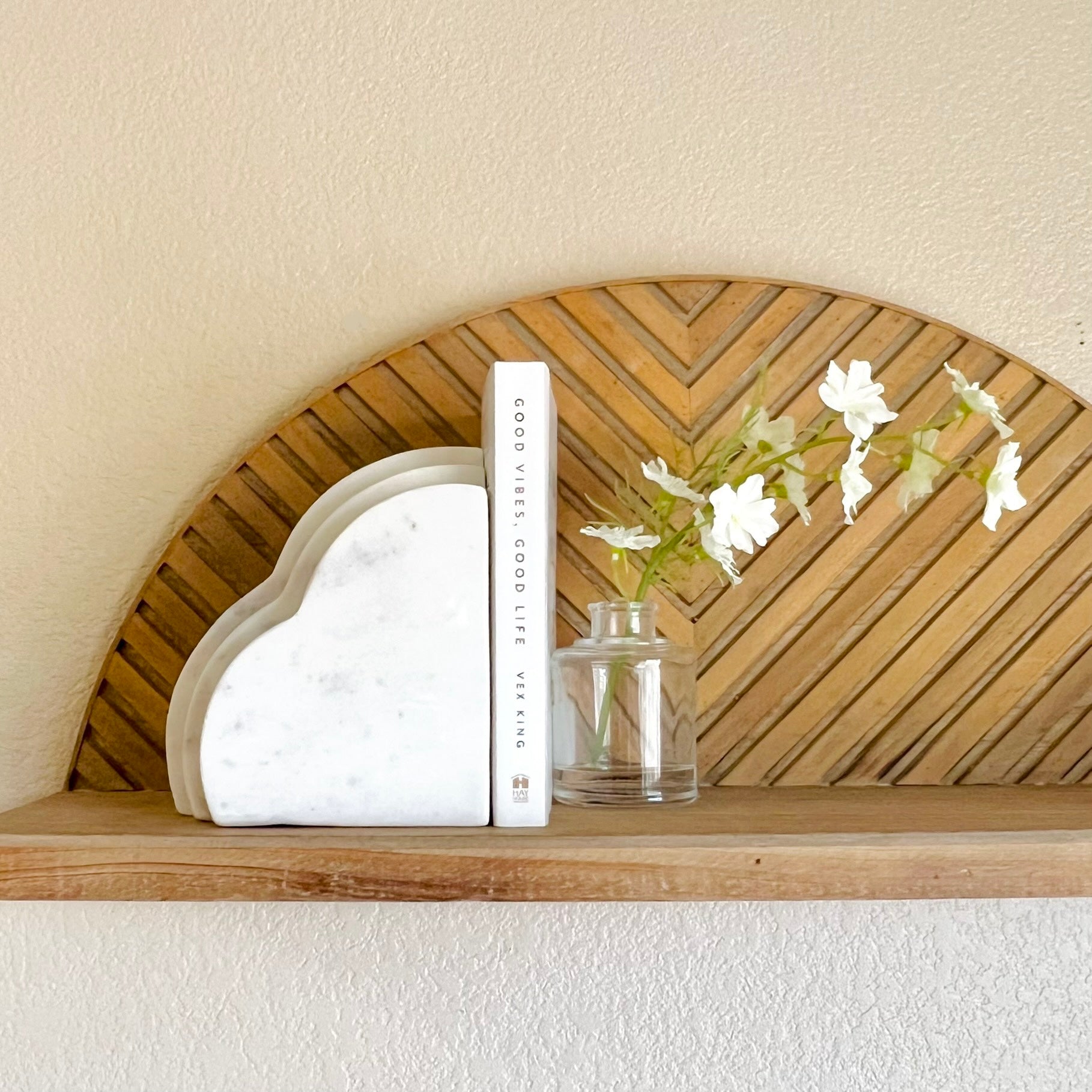 A white marble bookend shaped like a cloud on a wooden shelf with one white book and a small bud vase filled with little white flowers