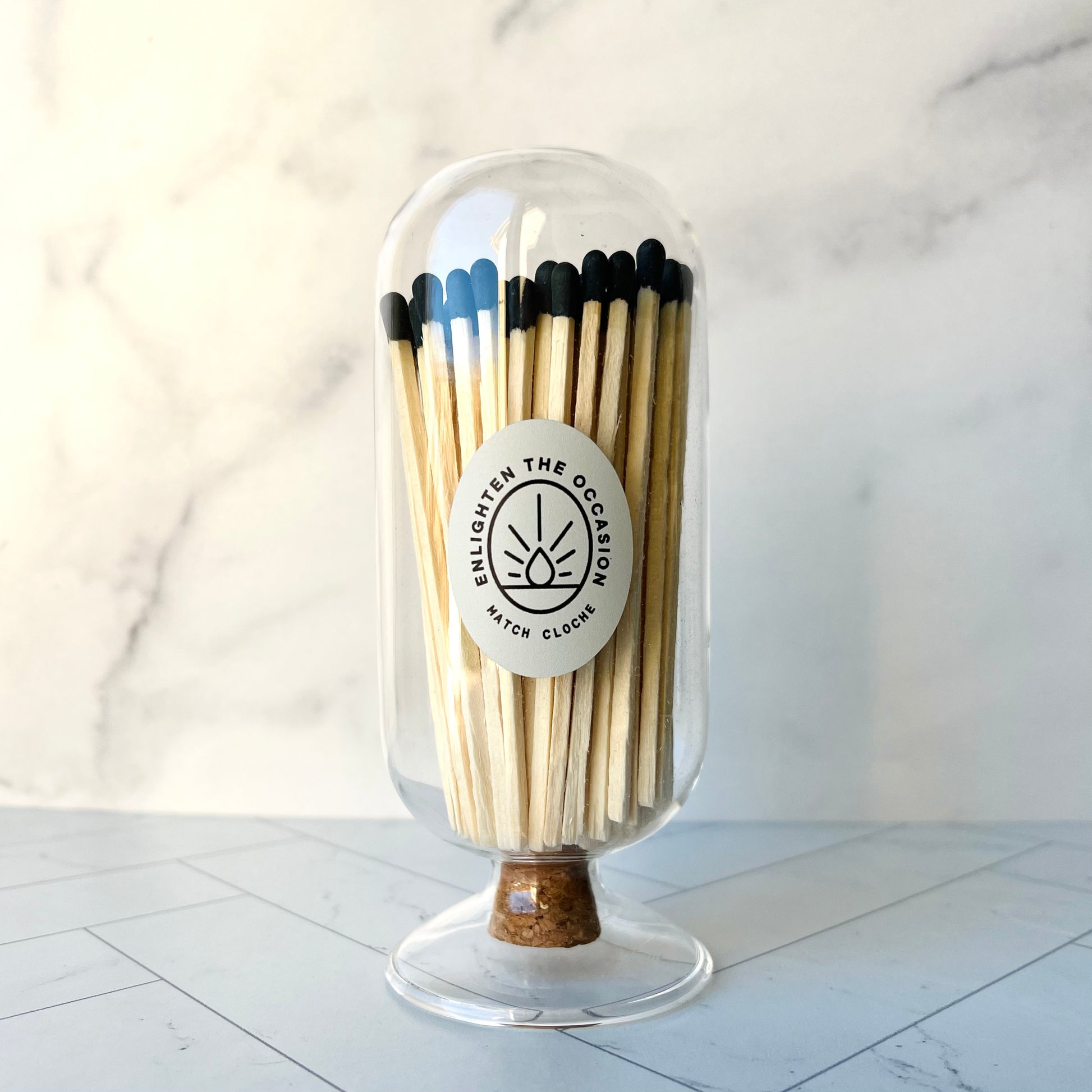 A clear glass cloche with a cork stopper on the bottom and filled with matches with black tips