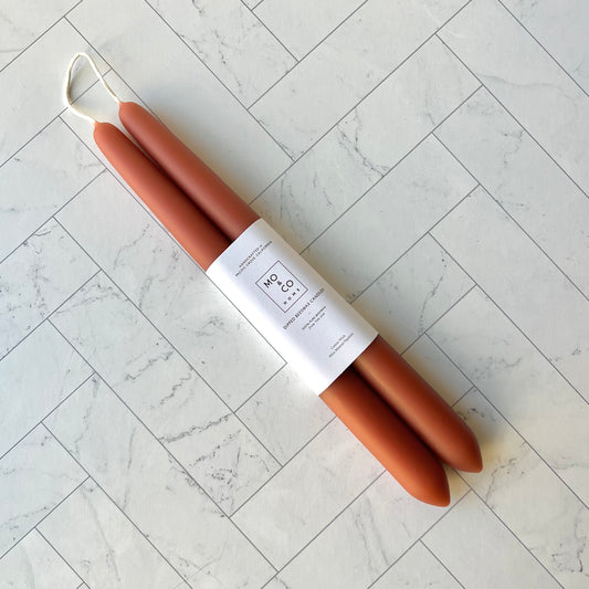 A set of two taper candles in a terracotta color lying diagonally against a white tiled surface