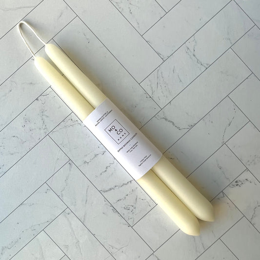 A set of two ivory white taper beeswax candles lying diagonally against a white tiled surface