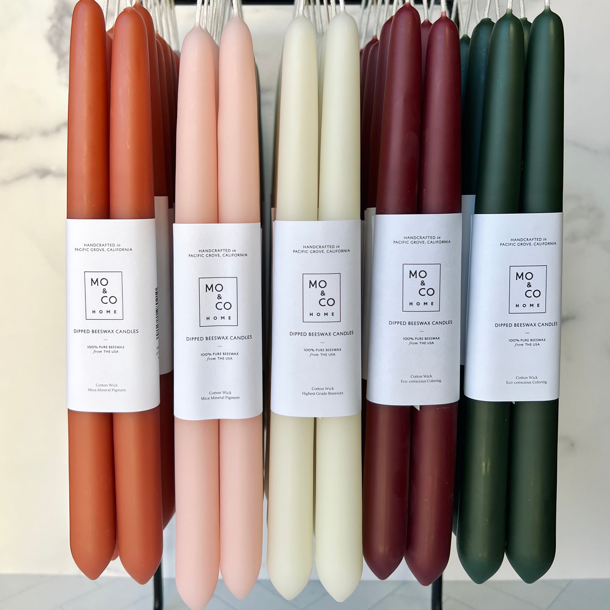 Sets of taper beeswax candles hanging from a display stand. From left to right they are in the colors: terracotta, light pink, white, burgundy and forest green