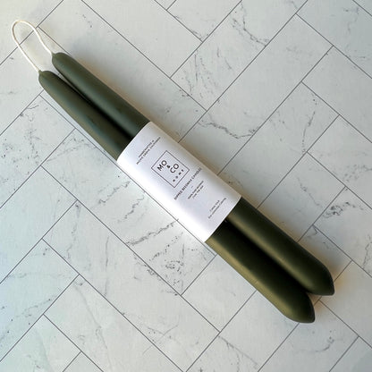 A set of two taper beeswax candles in forest green lying diagonally against a white tiled surface