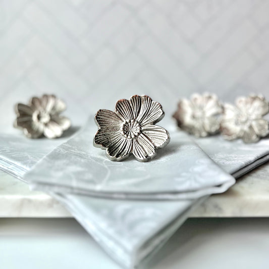 A silver flower napkin ring sitting on gray napkins with the other napkin rings in the set in the background