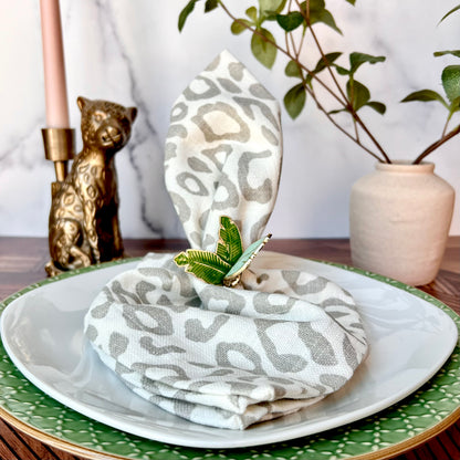A leopard print napkin shown folded with a napkin ring around in on a white plate with a plant in the background on the right and a leopard candlestick on the left