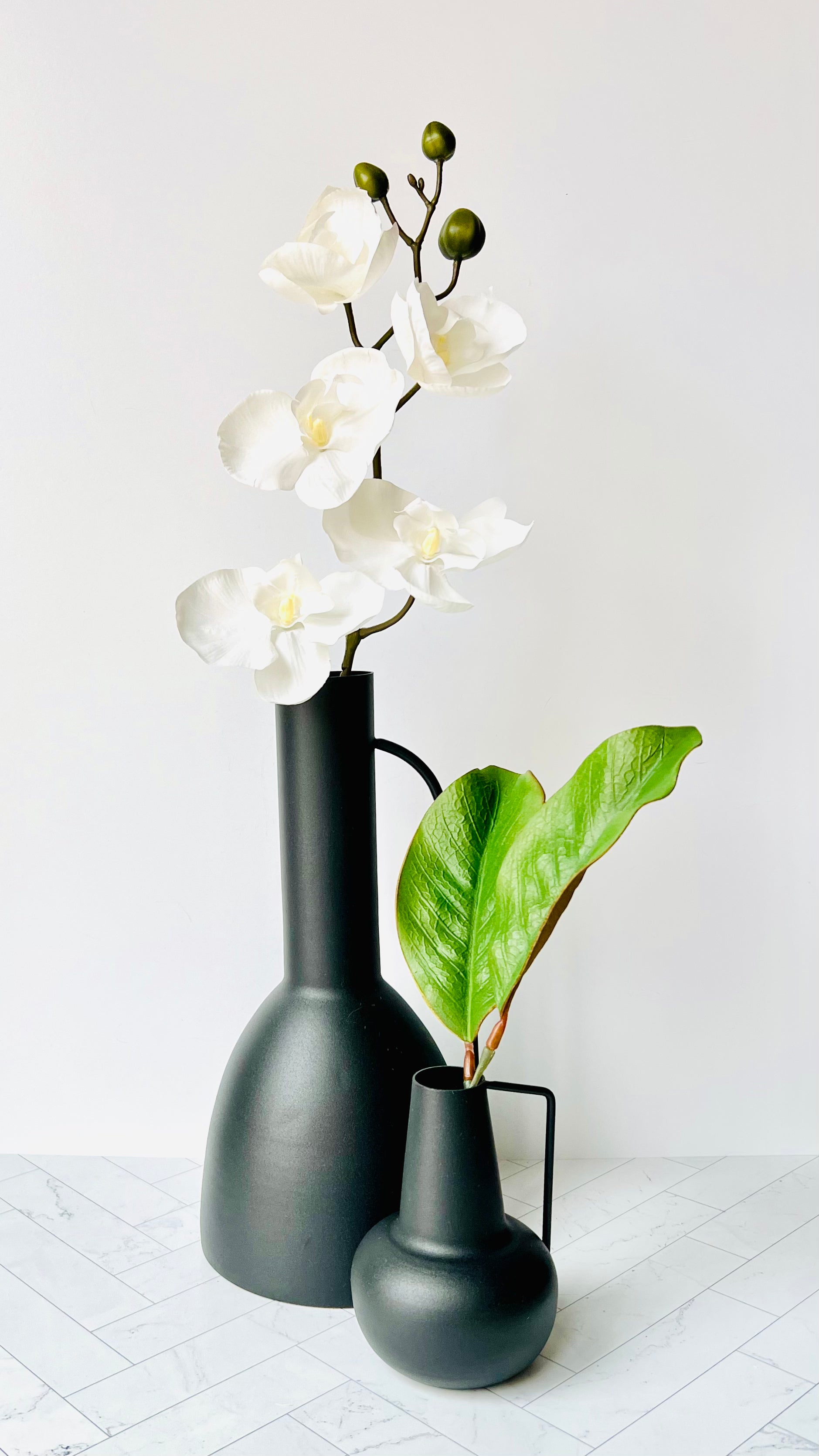 The Sleek Black Vase on a table filled with white flowers with the Sleek Bud Vase next to it filled with green leaves