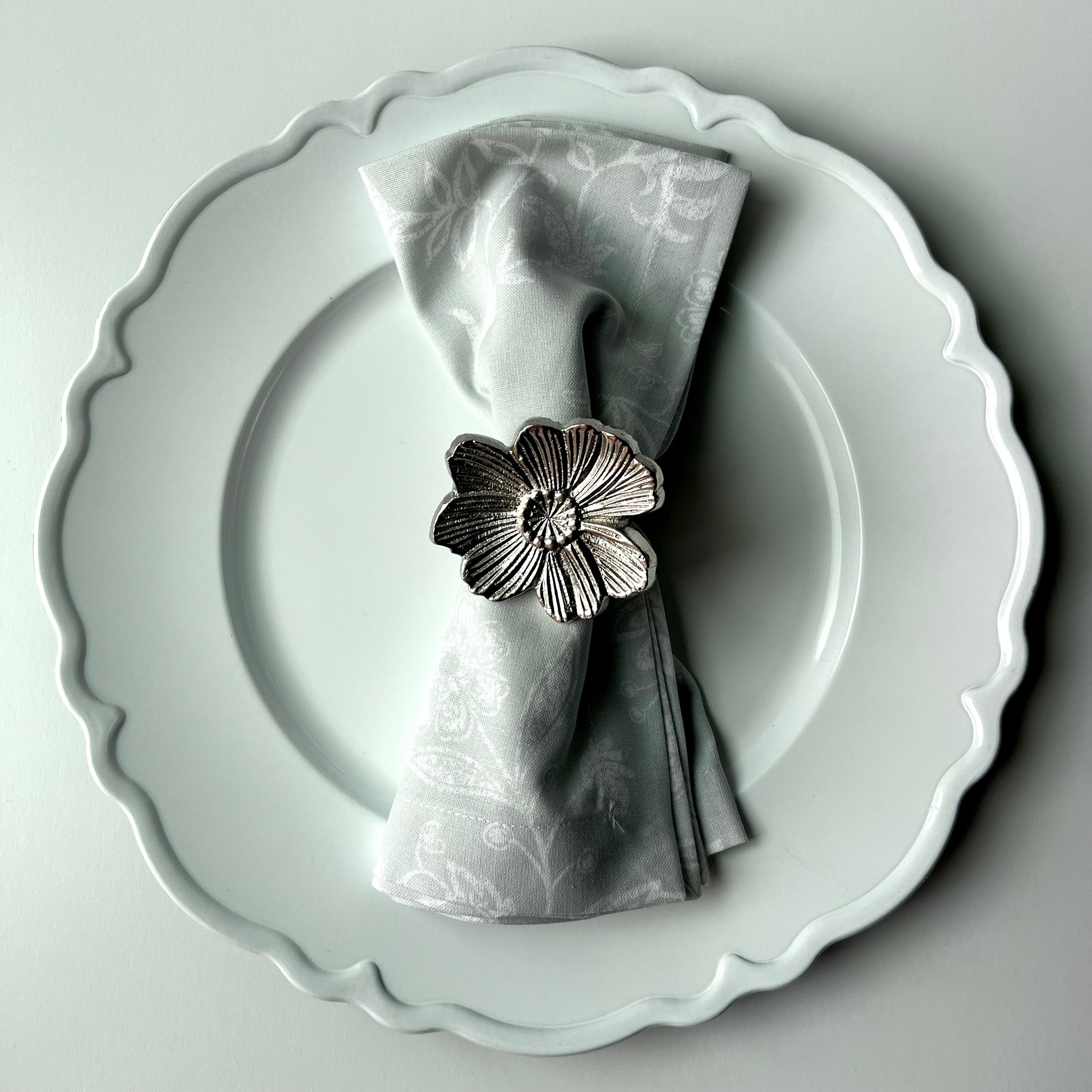 A silver flower napkin ring wrapped around a gray napkin on a white plate against a white background