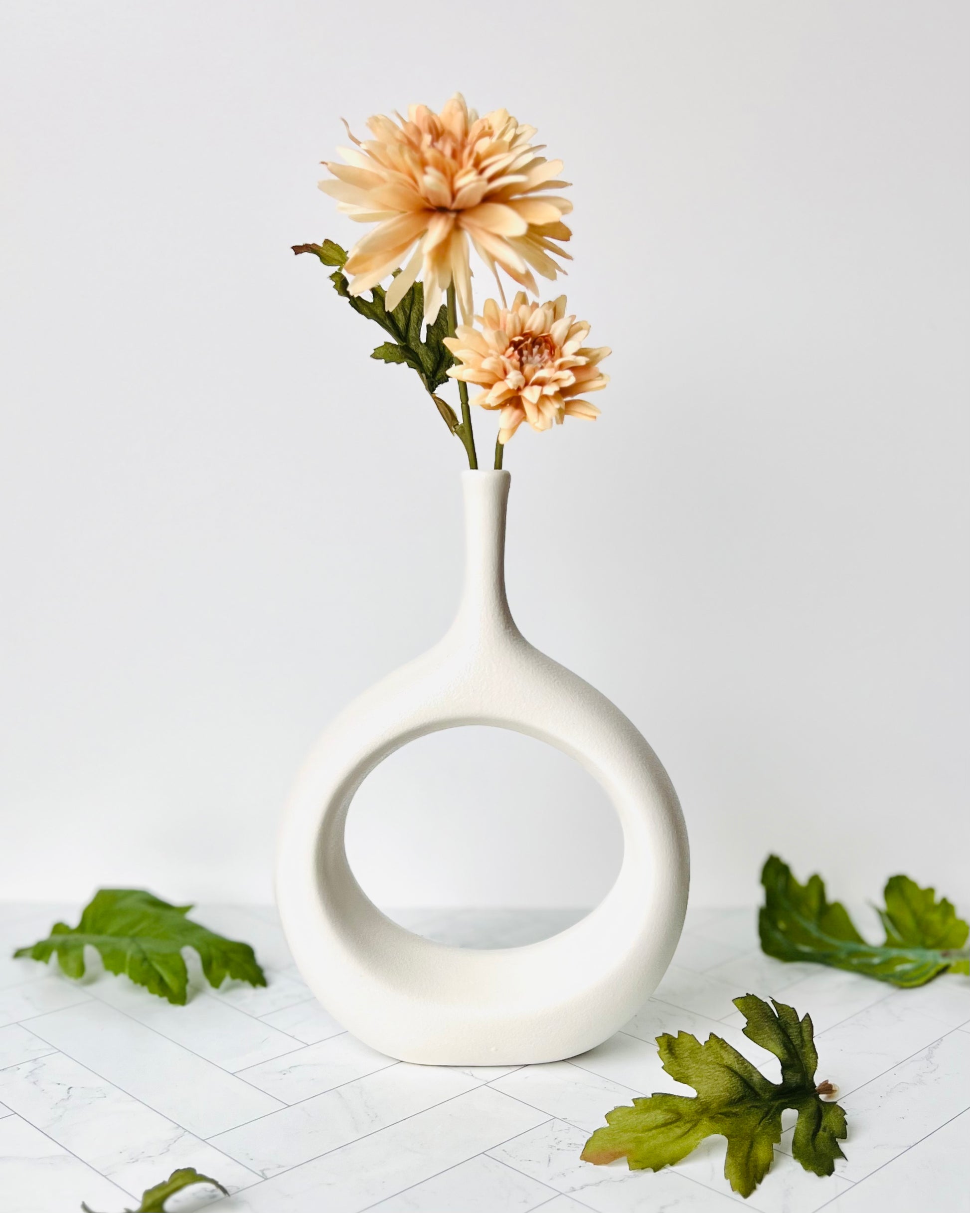 The Hollow Round Vase filled with an orange flower and with green leaves scattered around a white table