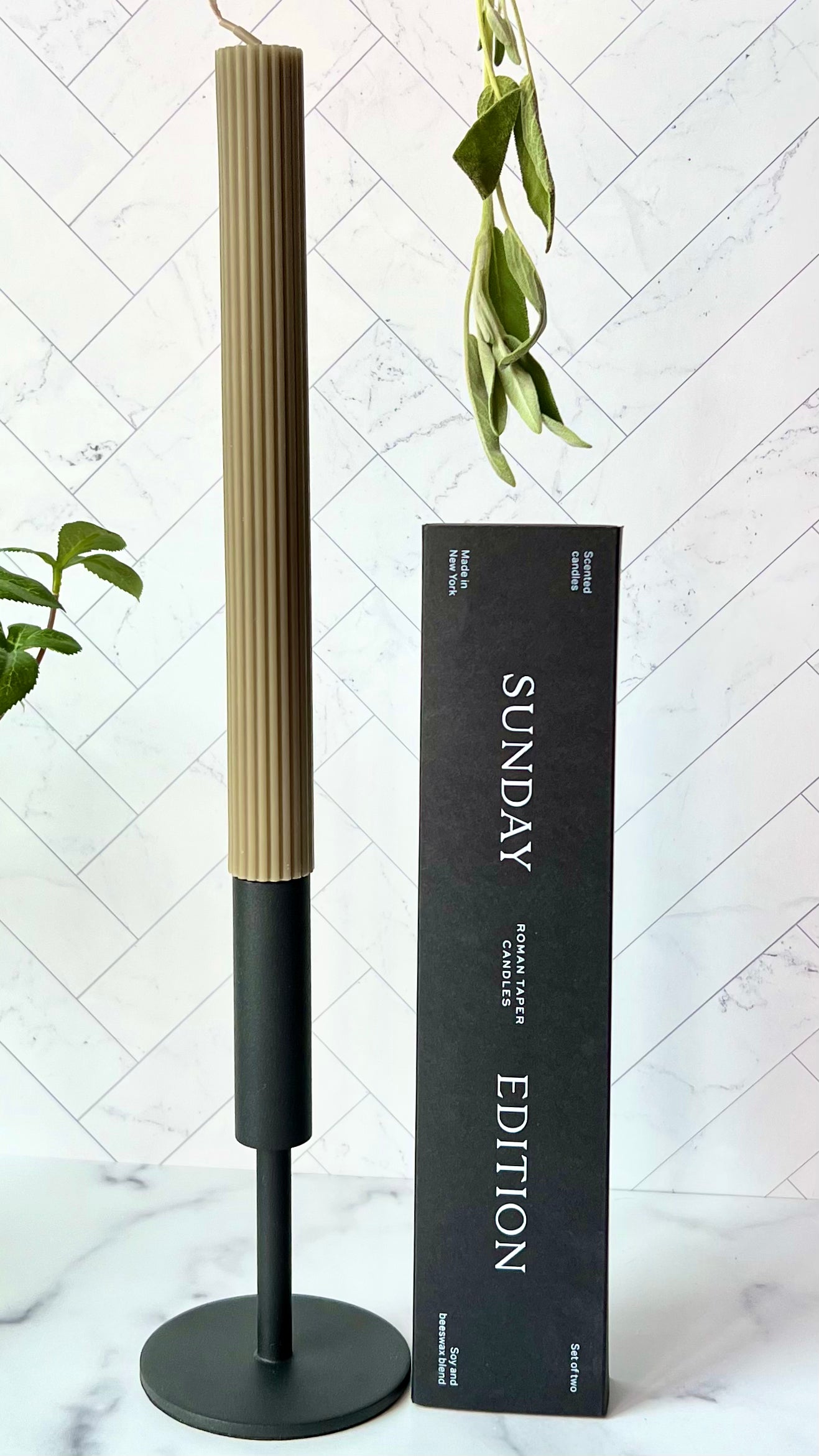 A sage green taper candle set in a black iron candlestick with its box set next to it and greenery around them