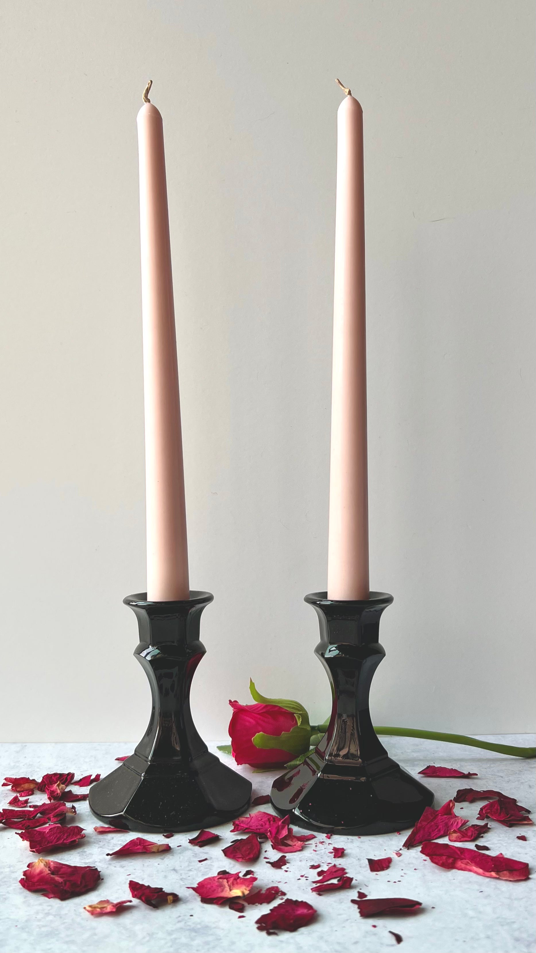 The Rose Taper Candles set in two black candlesticks set on a light gray surface that is scattered with red rose petals