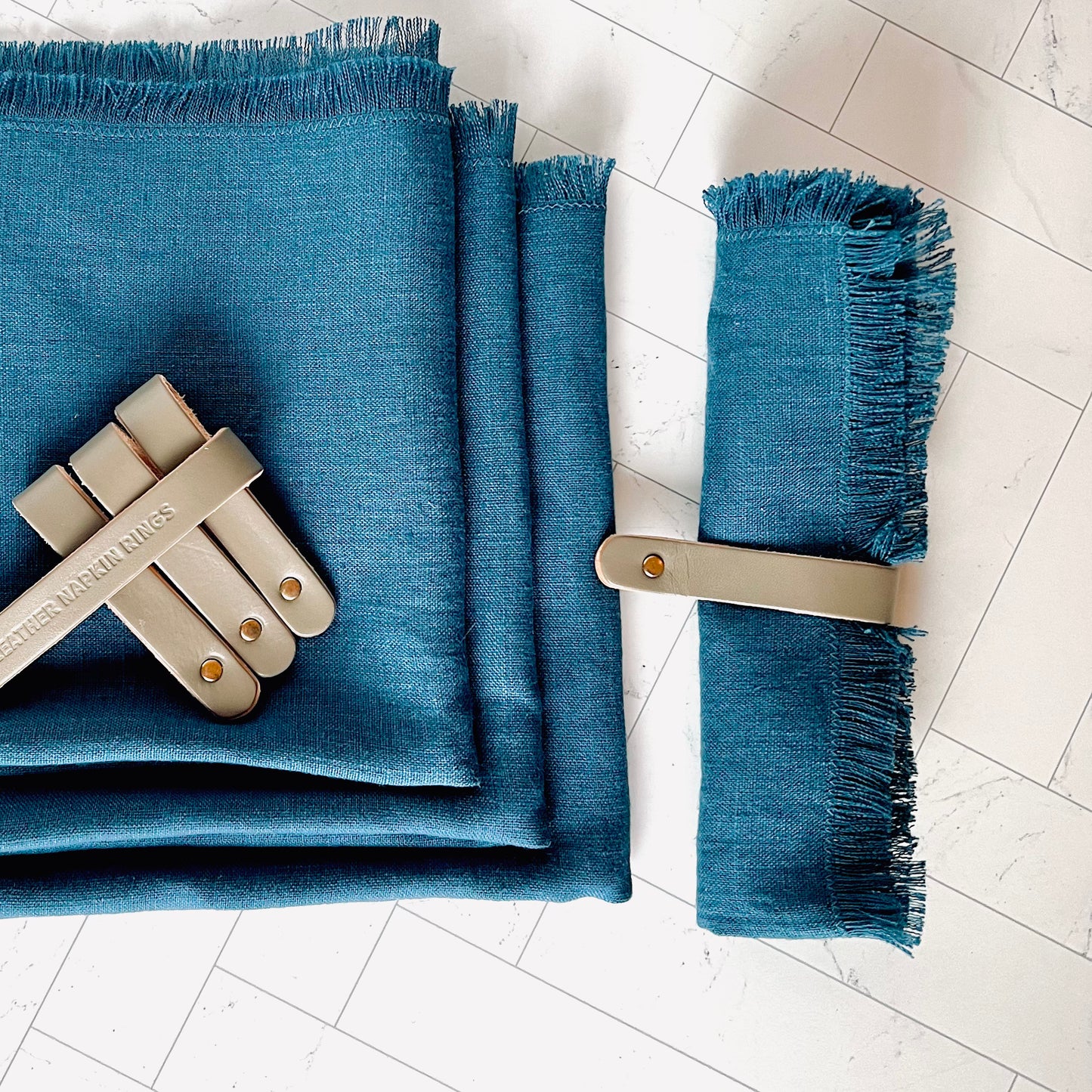 The Gray Leather Napkin Rings shown with one wrapped around a blue napkin and the others strapped together on top of a stack of blue napkins- The Offbeat Co.