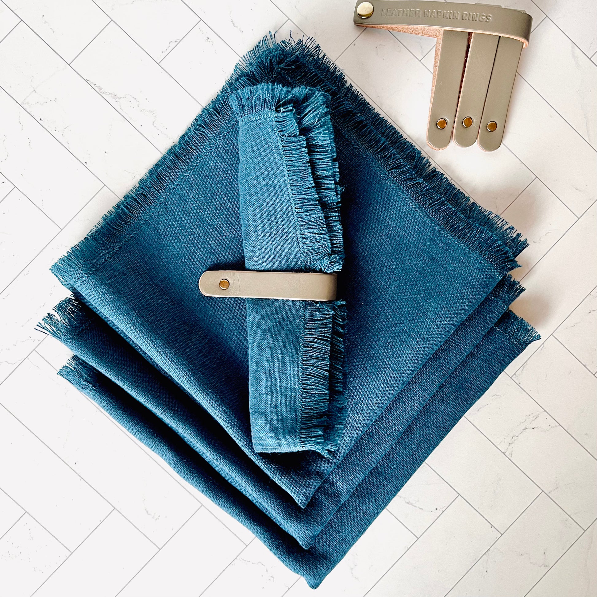 A Gray Leather Napkin Ring wrapped around a blue linen napkin - The Offbeat Co.