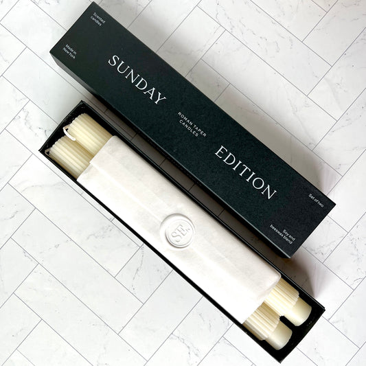 A set of two white taper candles in a black box with a white label wrapped around them
