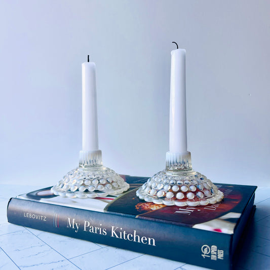 One white and clear hobnail candlestick with another in the background, both with taper candles inserted