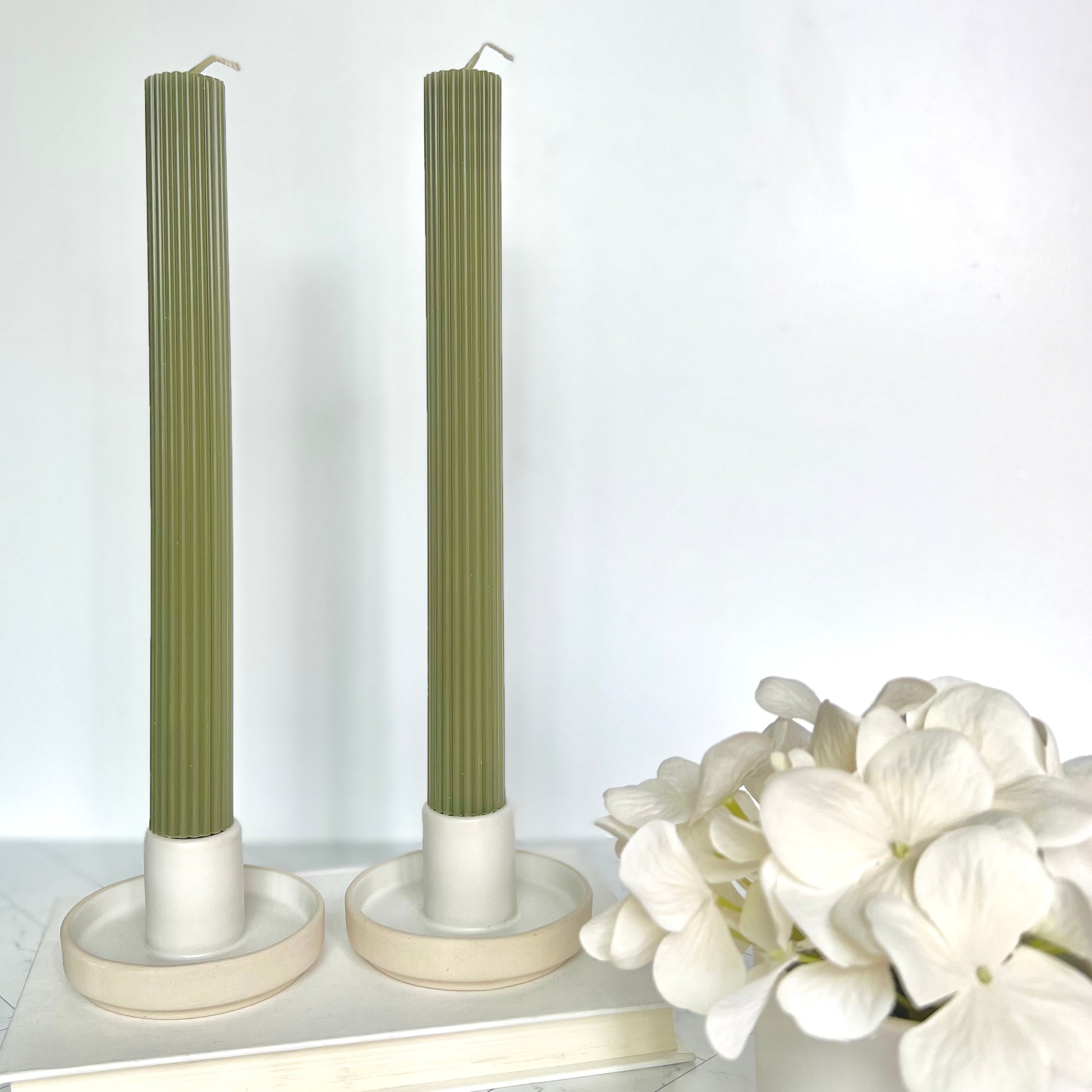 Two light green taper candles set in short, beige candlesticks with white flowers next to them