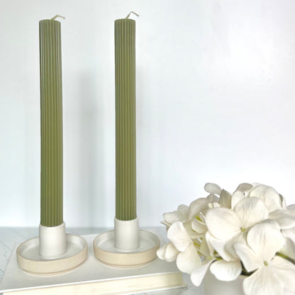 Two light green taper candles set in short, beige candlesticks with white flowers next to them