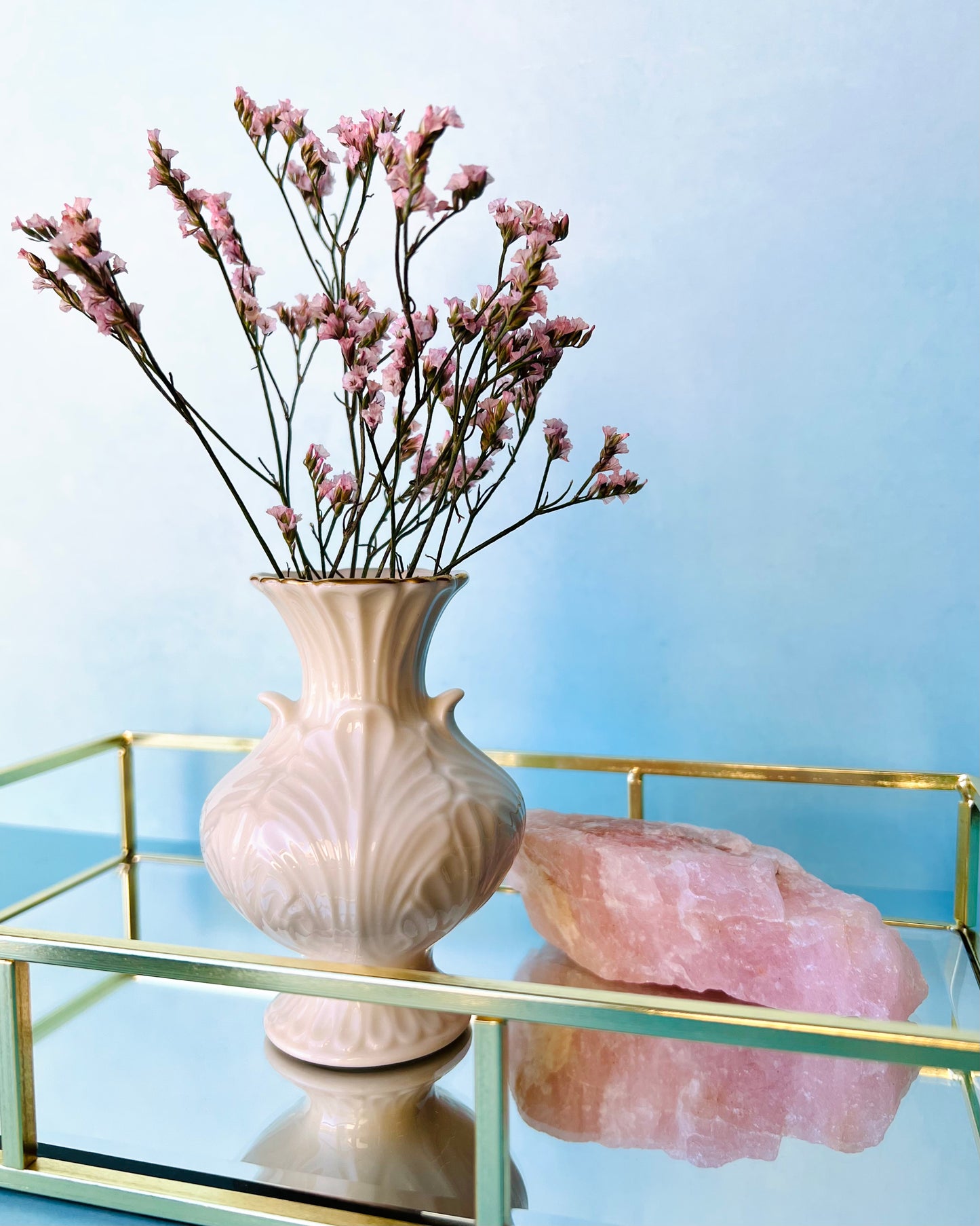 The vintage bud vase filled with sprigs of little pink flowers on a mirrored tray with gold edges with a pink stone behind