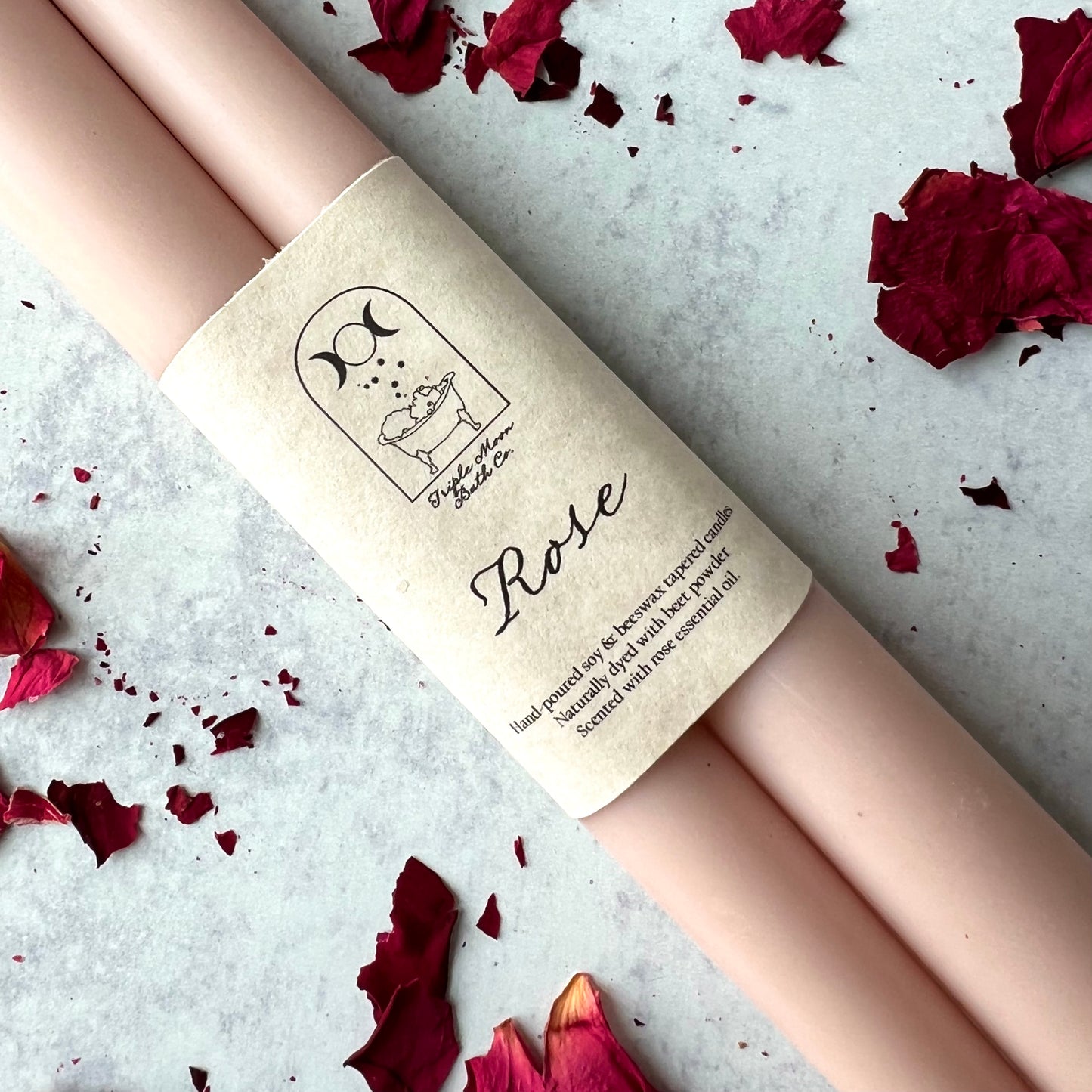 A close up of two light pink taper candles wrapped in a beige wrapper with black writing and rose petals scattered around them