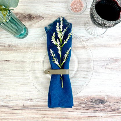 A dark blue linen napkin wrapped in a gray leather napkin ring set against a light wooden surface with a sprig of greenery tucked into the napkin ring with a glass of wine on the top of left and more greenery in the top left