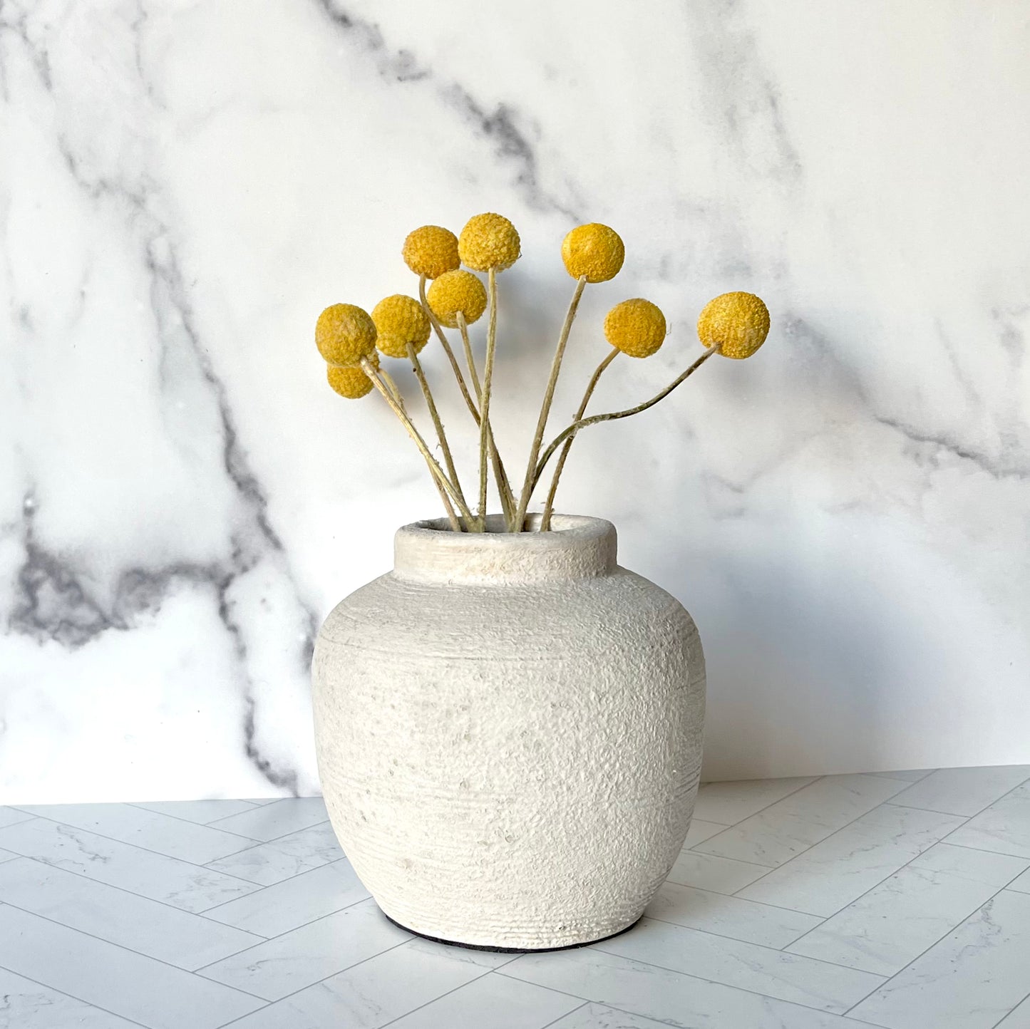 A small light gray vase filled with yellow dried billy button flowers against a white marble wall