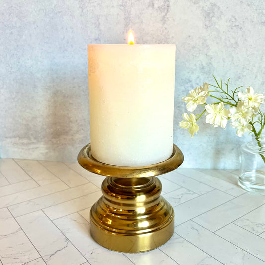The Vintage Pillar Candlestick with a large white candle set on it and a small cluster of white flowers in the background - The Offbeat Co.