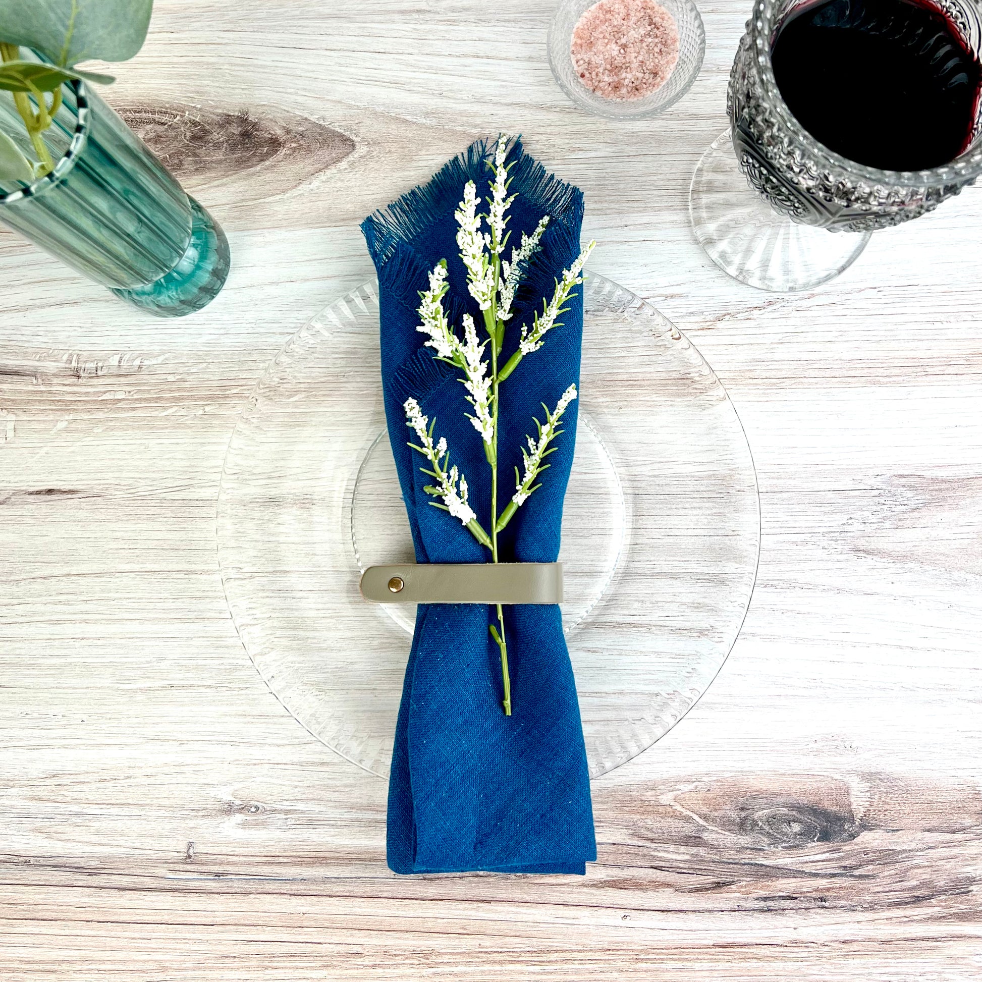 A Gray Leather Napkin Ring shown overhead wrapping a blue napkin with a sprig of greenery tucked inside- The Offbeat Co.