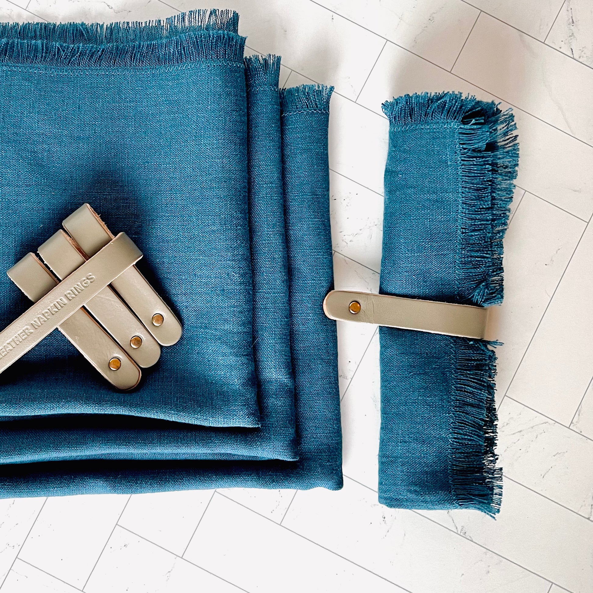 A blue linen napkin wrapped in a gray leather napkin ring with a three other napkins and rings on the side