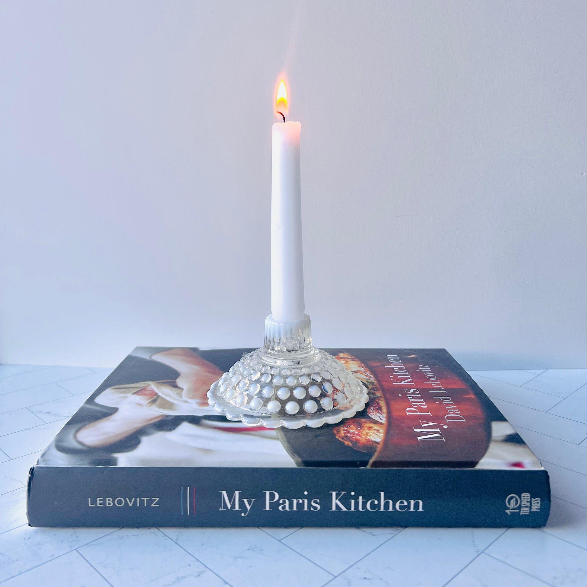 One white and clear hobnail candlestick holding a lit candle on a black cookbook